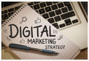 Hand-drawn text reading "DIGITAL MARKETING STRATEGY" on a lined spiral notebook, accompanied by doodles of a magnifying glass, thumbs up, emails, and arrows. A blue pen rests on the notebook, which is placed on a laptop keyboard.